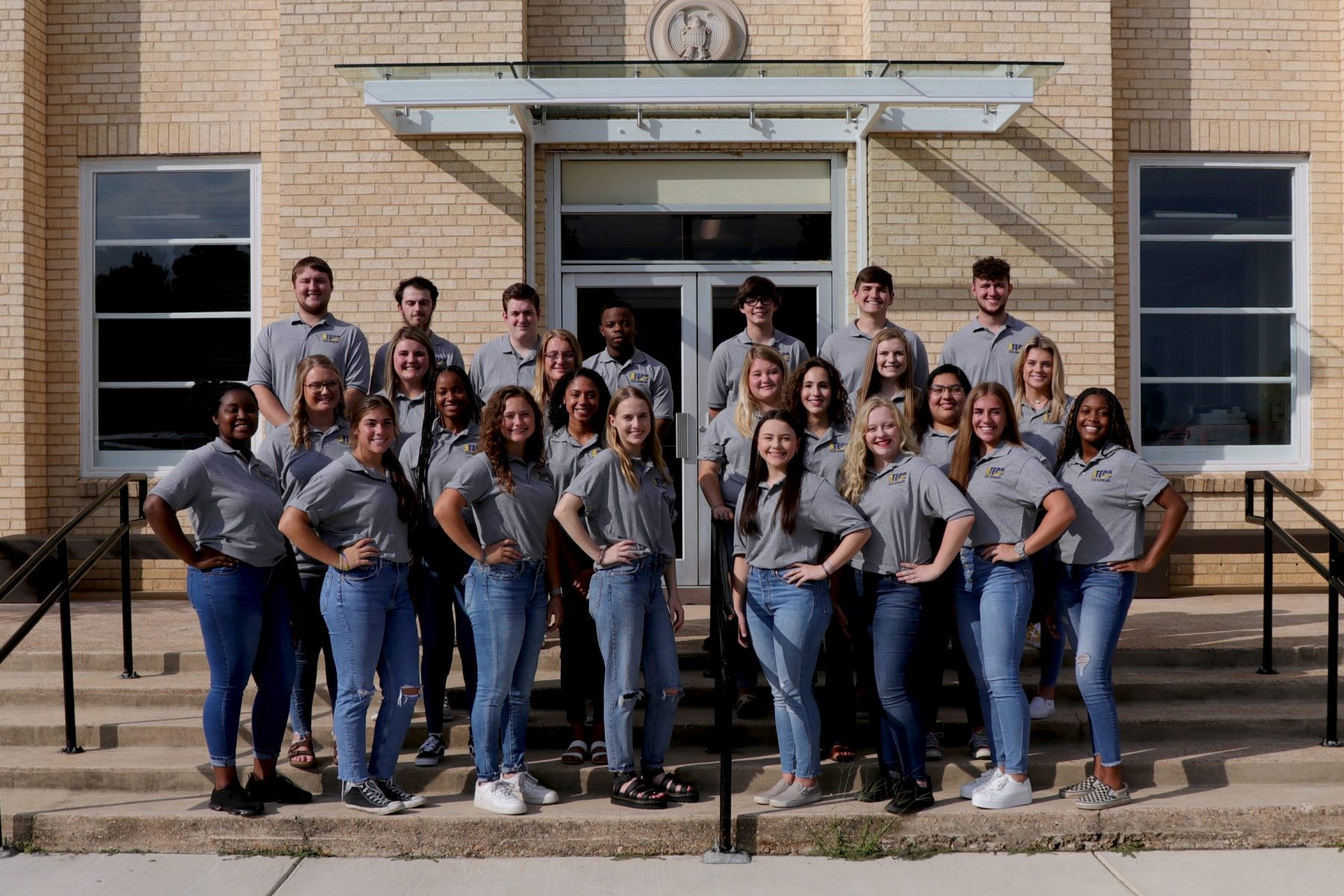 students in gray shirts and jeans on the front steps of a brick building
