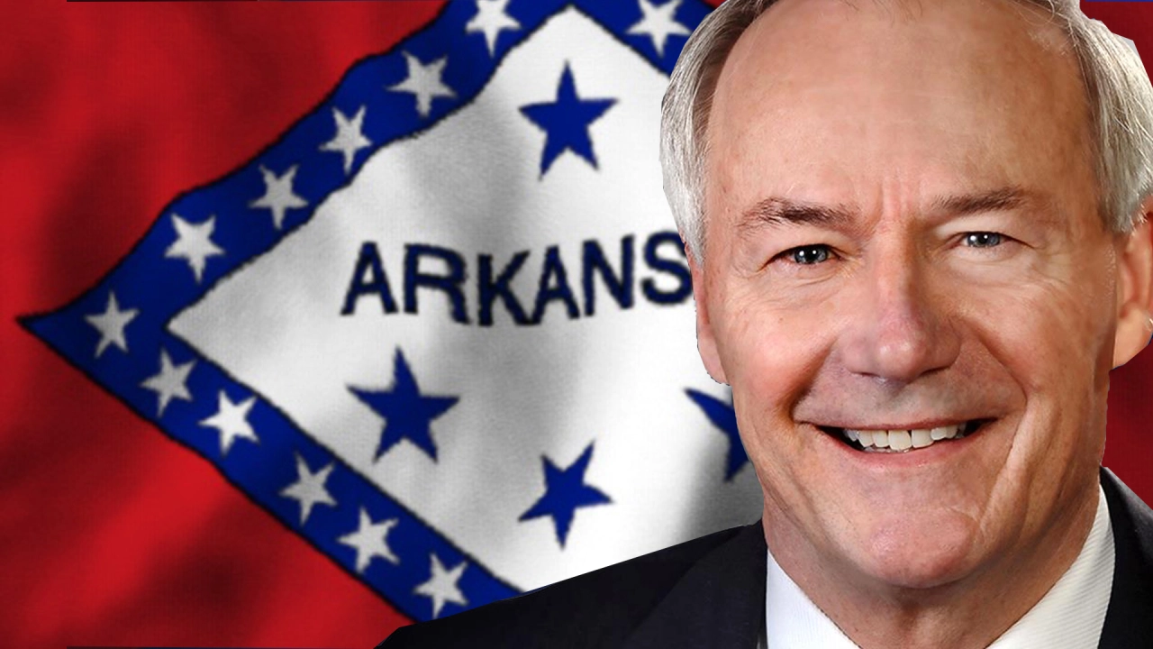man in a suit smiling with the Arkansas flag behind him